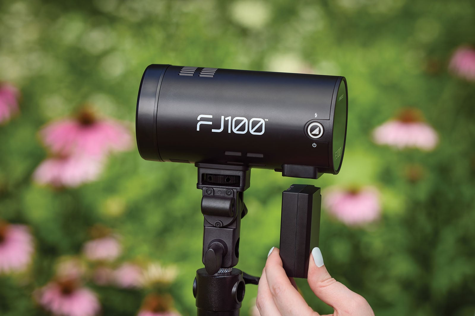 A hand holds a black battery pack near the slot of a black FJ100 flash unit mounted on a light stand against a blurred floral background.