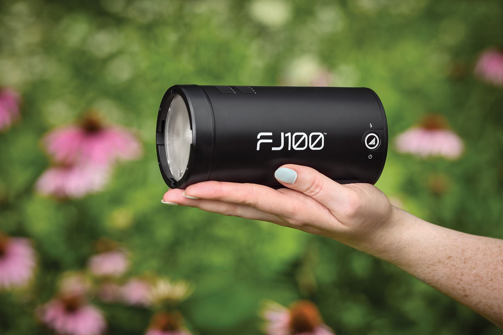 A person's hand holds a black, cylindrical FJ100 portable flash unit against a blurred background of pink flowers and green foliage.