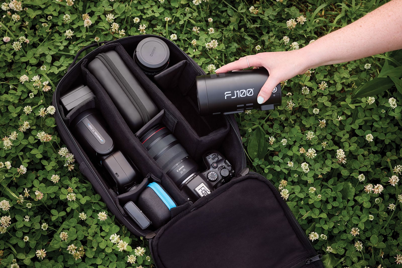 A hand places a Westcott FJ100 strobe light into a black camera bag filled with photography gear, including a Canon camera and lens, resting on a patch of clover.