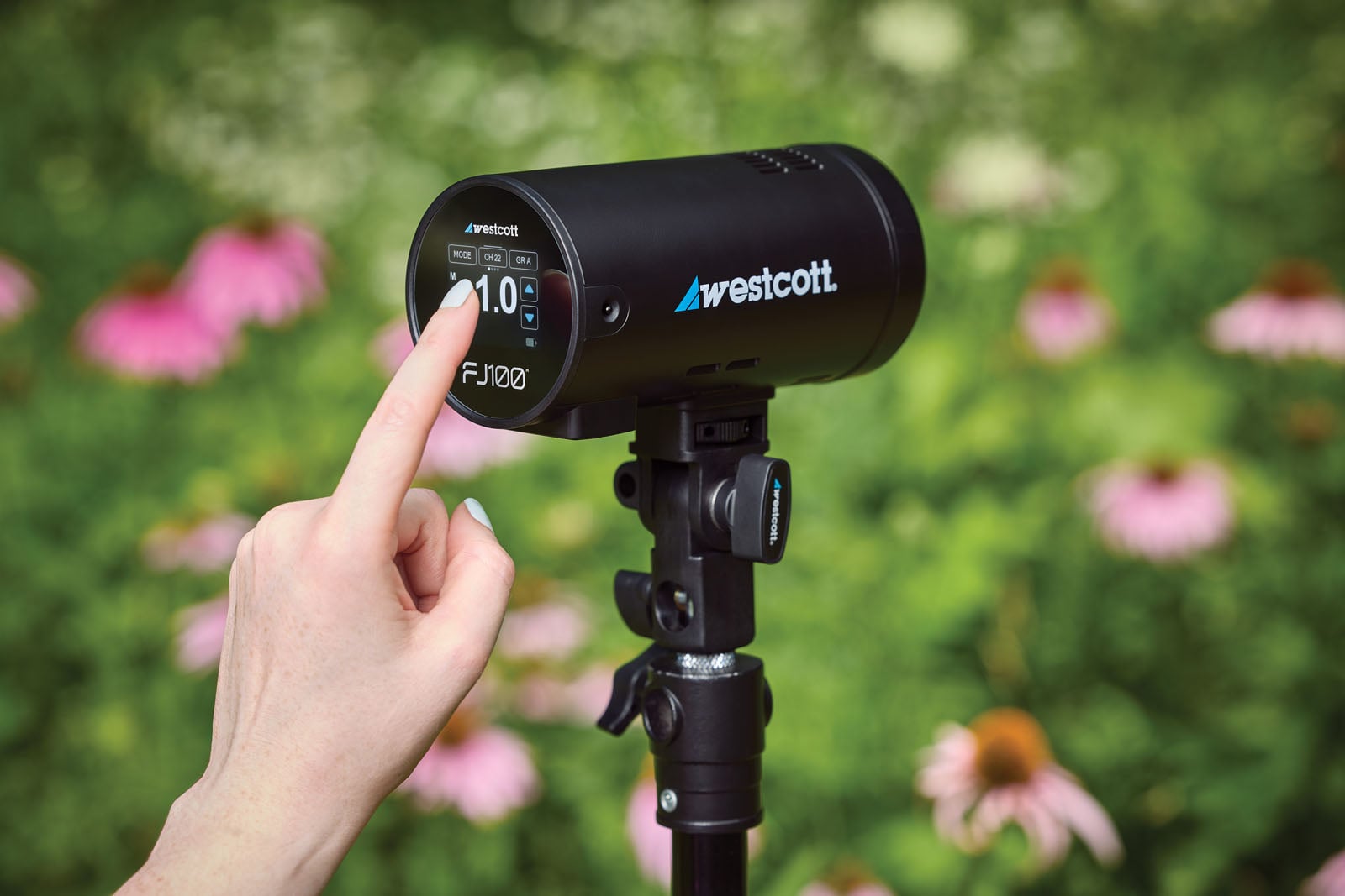 A hand touches the digital display of a black Westcott FJ100 strobe light mounted on a stand in a garden with pink flowers.