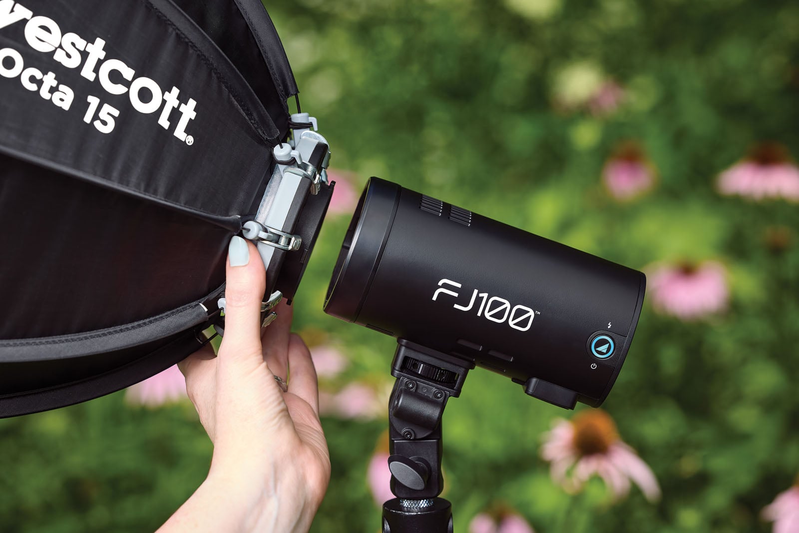 A person's hand holds a Westcott Octa 15 softbox near an FJ100 strobe light mounted on a stand in front of a blurred garden background.