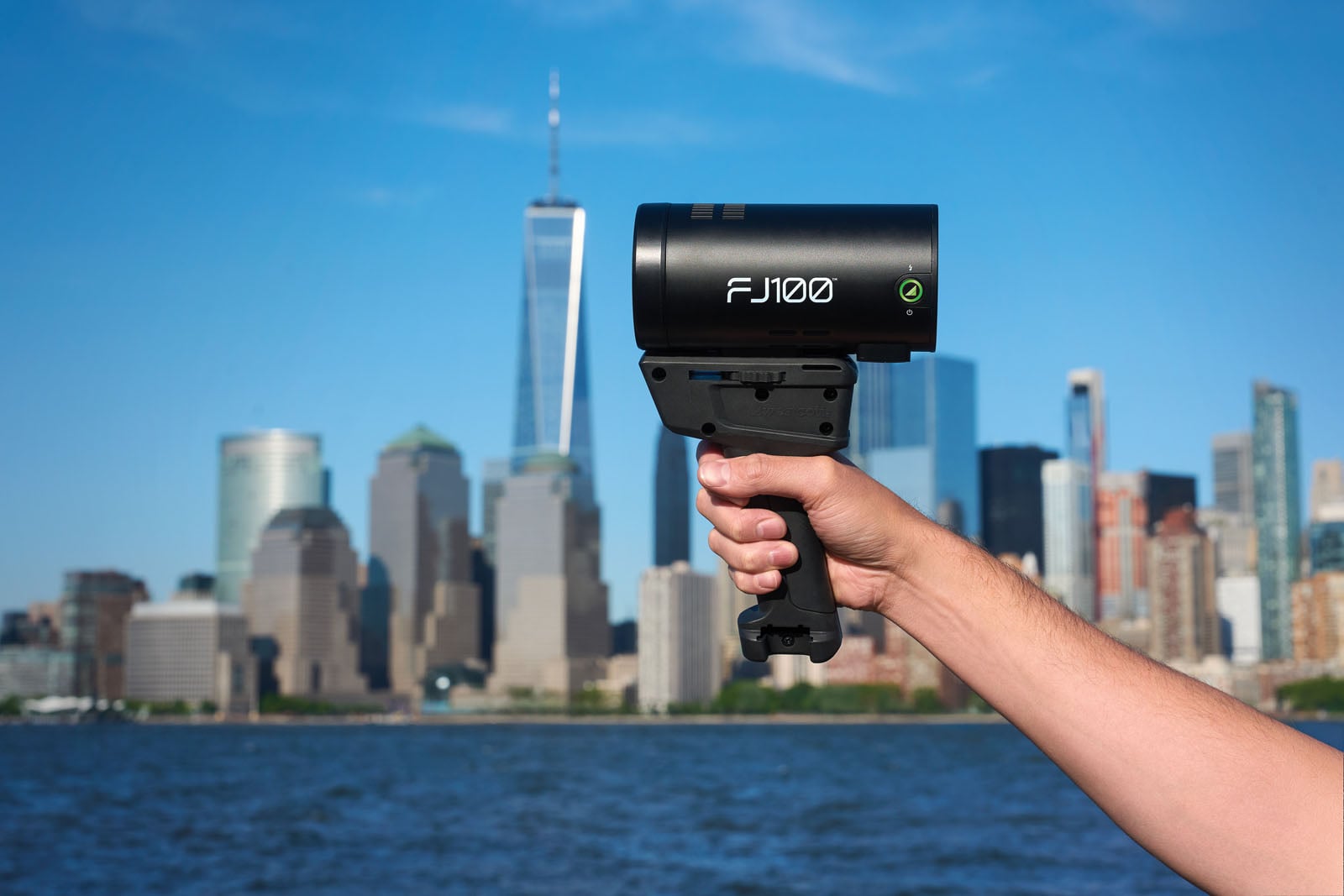 A hand holds a black FJ100 strobe light against a background of a city skyline and a body of water.