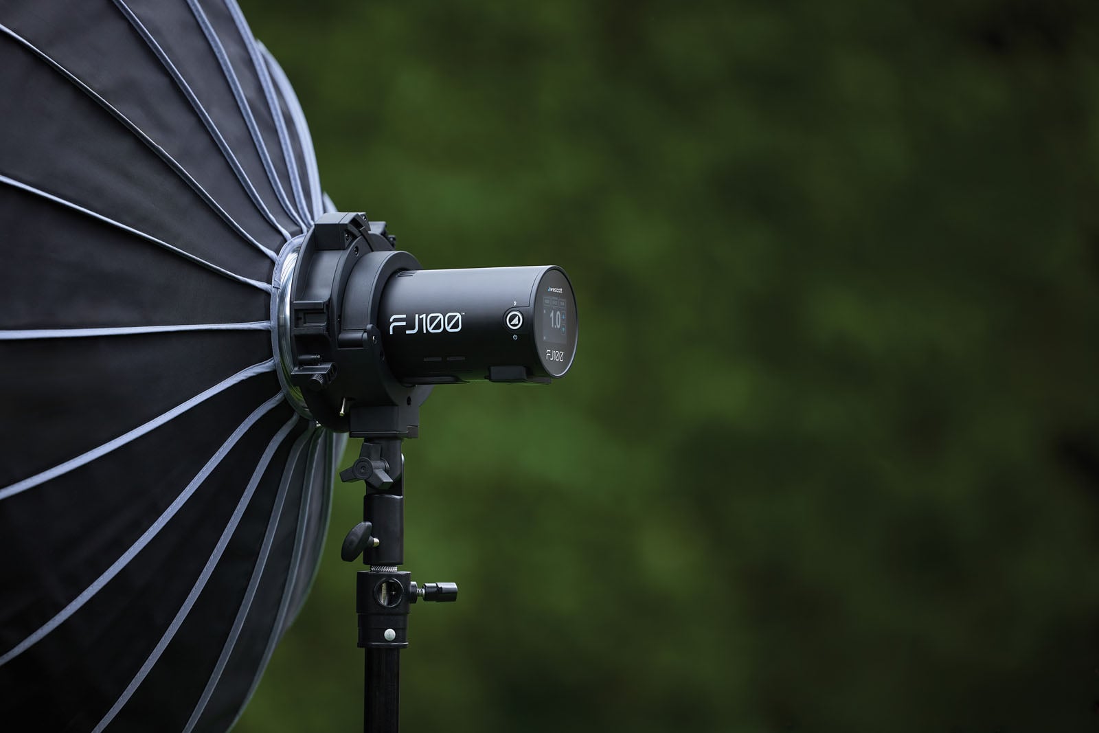 A Westcott FJ100 strobe light with a large softbox attached, mounted on a black light stand against a blurred green background.