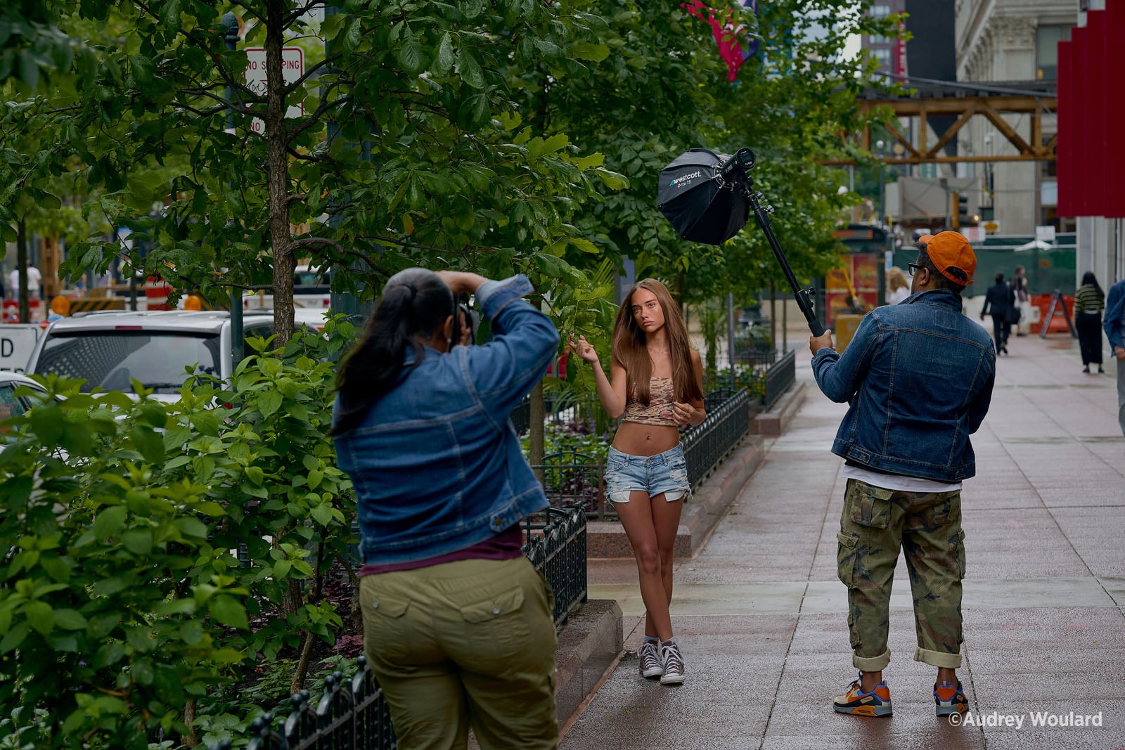 A photographer and an assistant holding a light modifier conduct a photoshoot with a young woman standing on a city sidewalk.