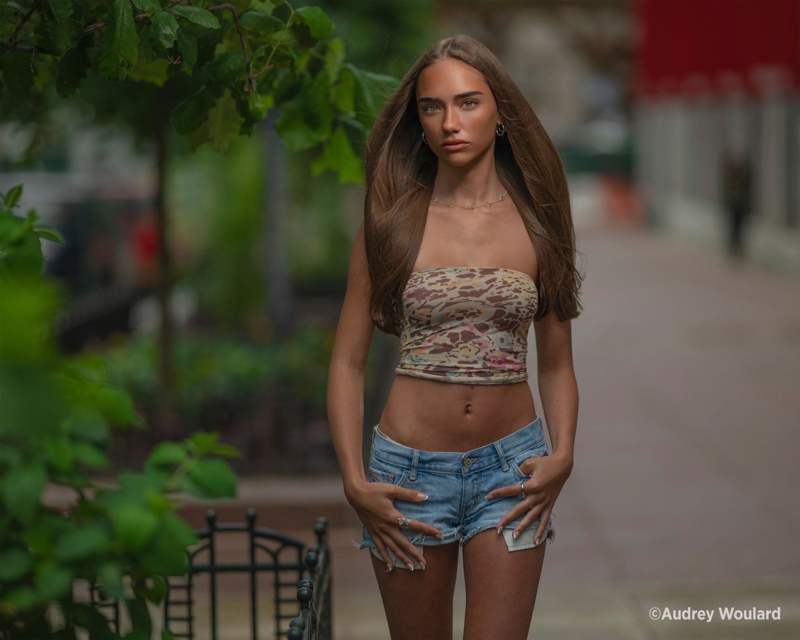 A young woman with long brown hair stands outdoors wearing a patterned tube top and denim shorts, with her hands tucked into her pockets.