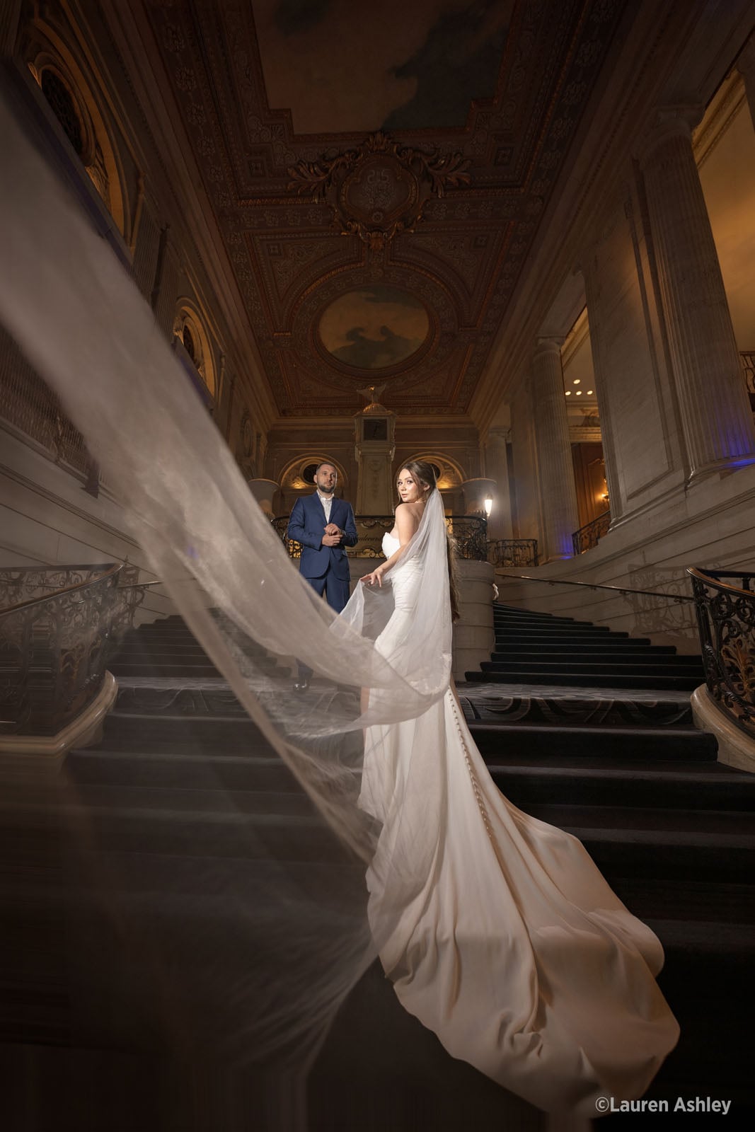 A bride in a white gown with a long, flowing veil stands on a grand staircase, looking back toward a man in a blue suit.