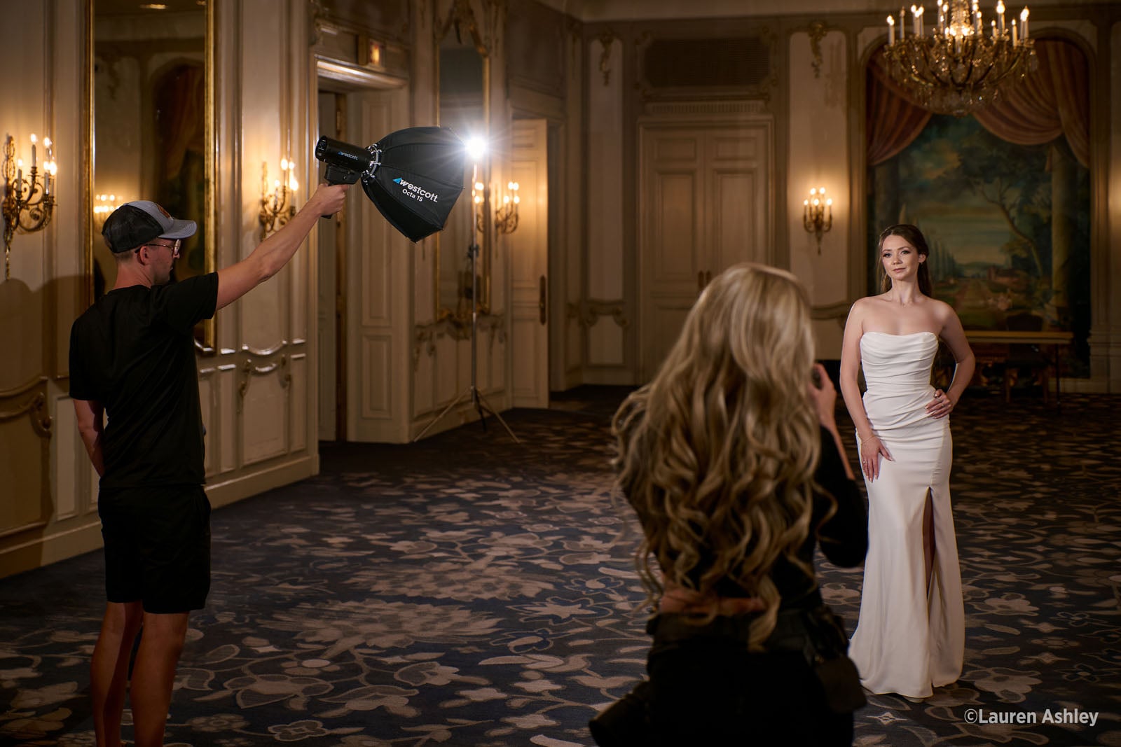 A photographer captures a portrait of a woman in a white gown while an assistant holds a portable light above her.