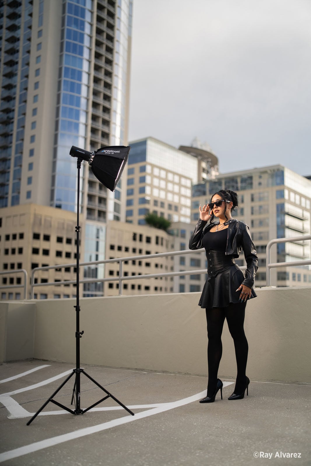 A woman in a black leather jacket, corset, and skirt stands on a rooftop parking deck next to a professional photography light on a stand.