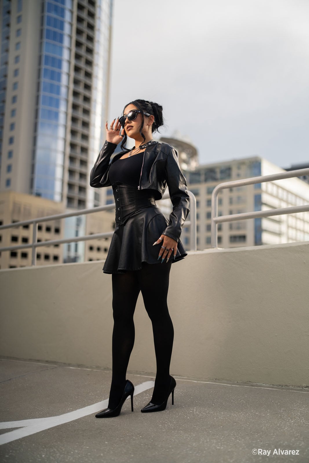 A woman in a black leather jacket, corset, and skirt with black tights and heels stands on a rooftop parking deck.