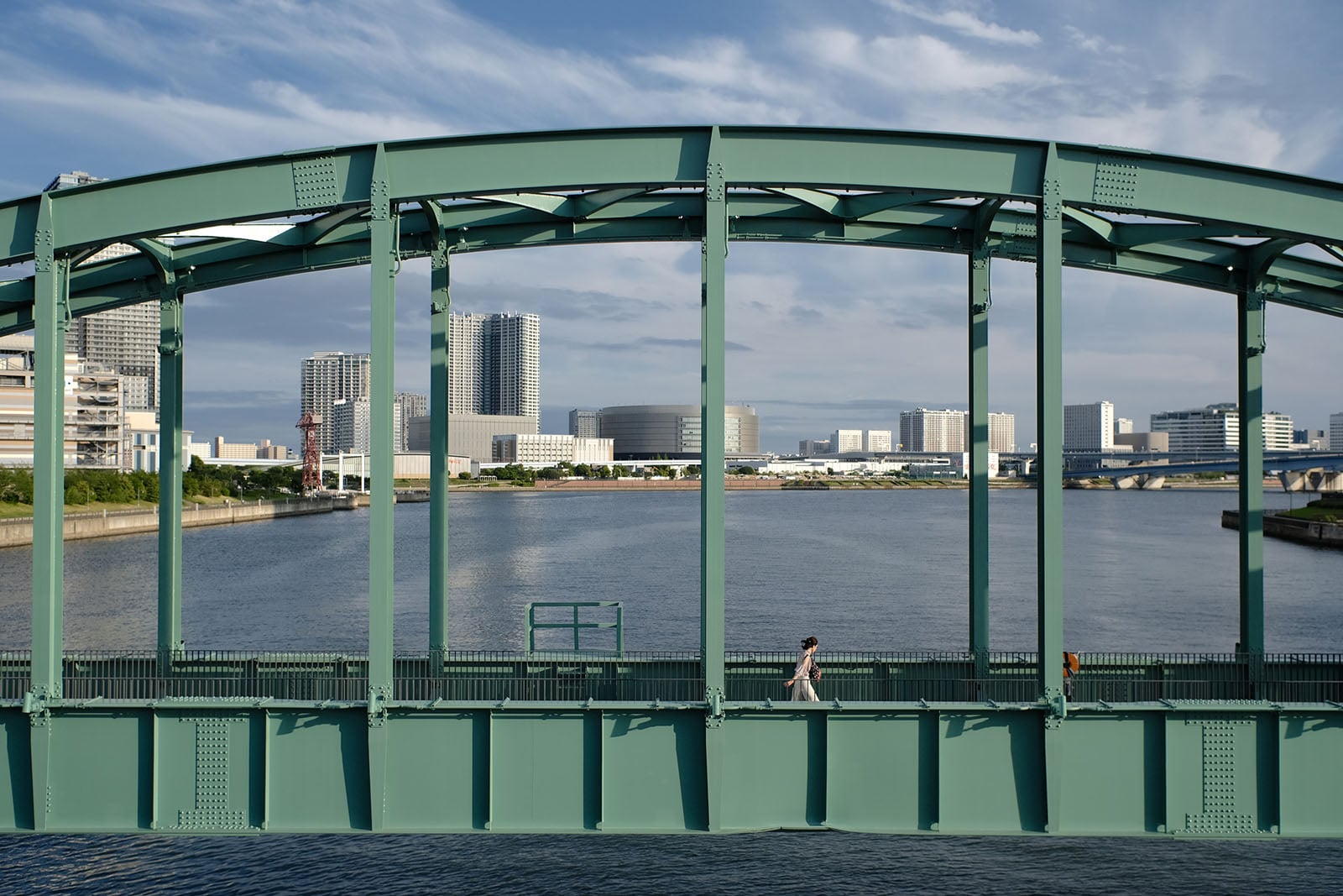 A person walks across a green metal truss bridge over a river with a city skyline in the background.