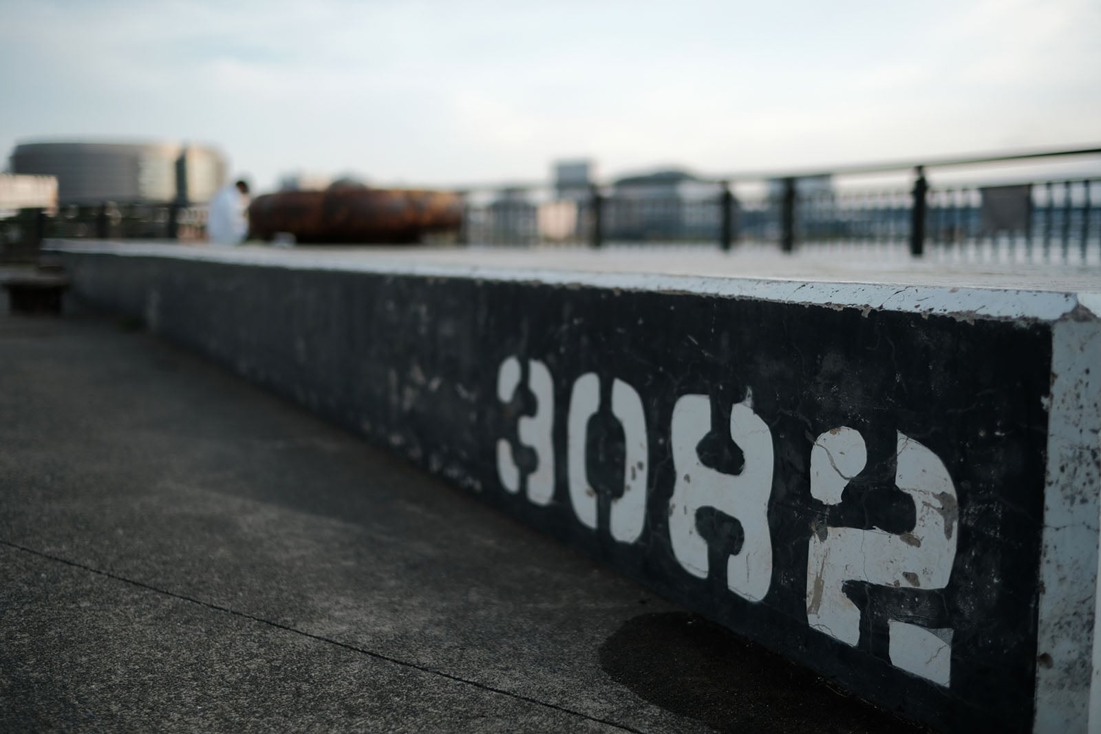 A close-up of a low concrete wall painted black with the white stenciled numbers "3082" on its side, with a blurred waterfront background.