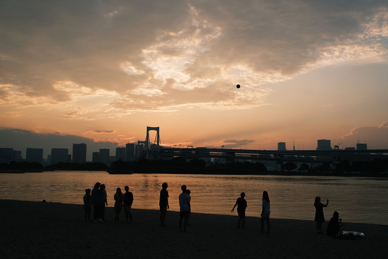 Silhouetted people stand on a beach at sunset with a bridge and city skyline in the distance, as a ball floats in the air.