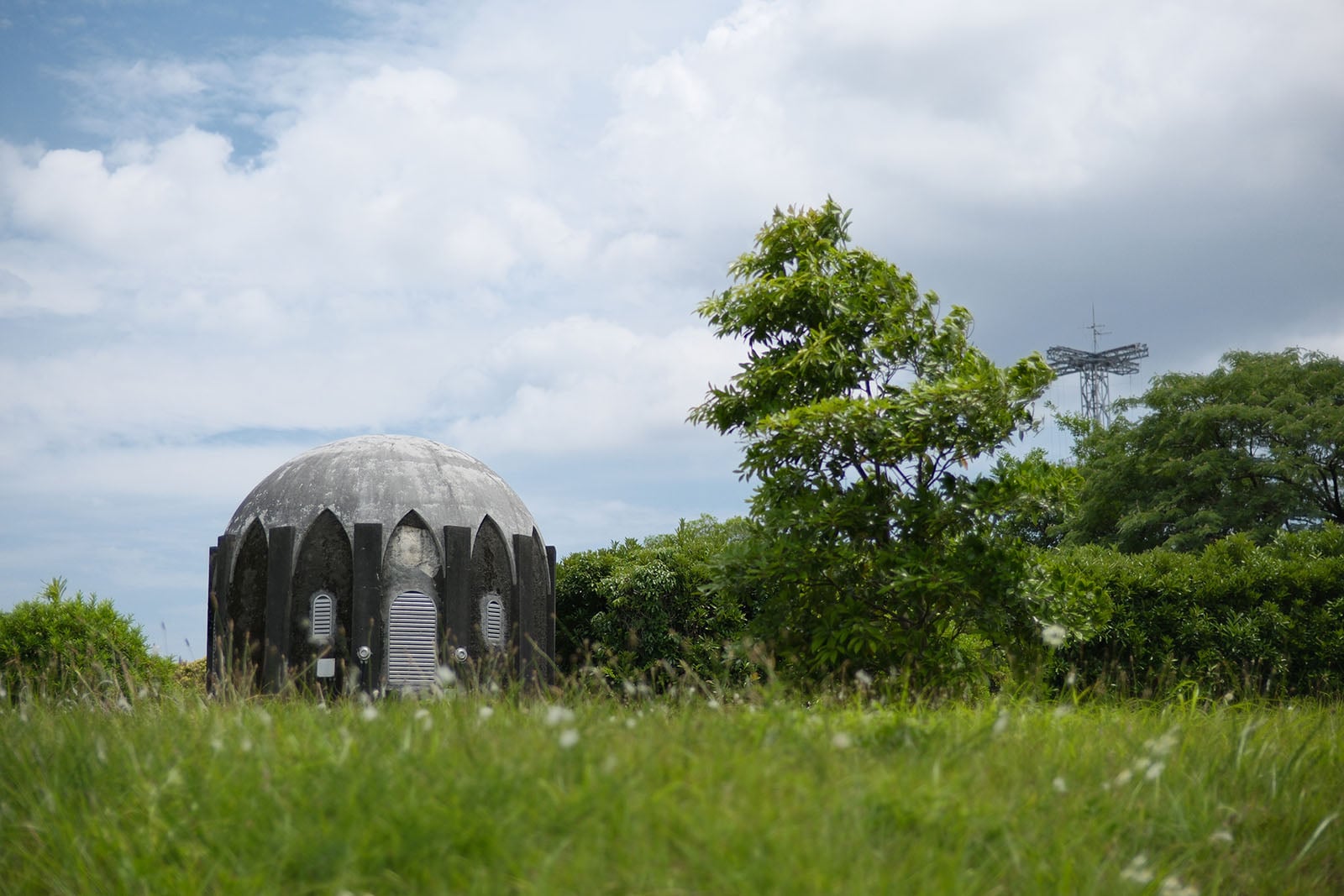 A concrete dome-shaped structure with arched panels sits in a grassy field next to a tree and a distant radio tower.