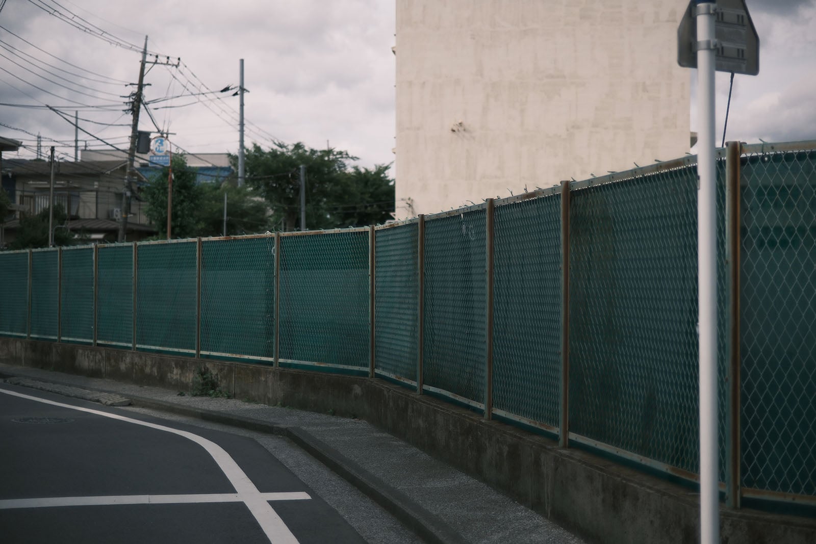 A green chain-link fence runs along a concrete wall beside a curved asphalt road under a cloudy sky.