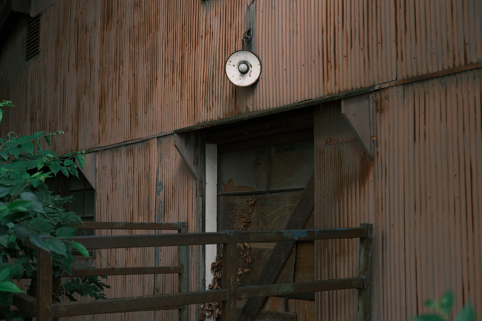 A rusted corrugated metal wall features a small, circular light fixture above a doorway partially obscured by a metal railing and green foliage.
