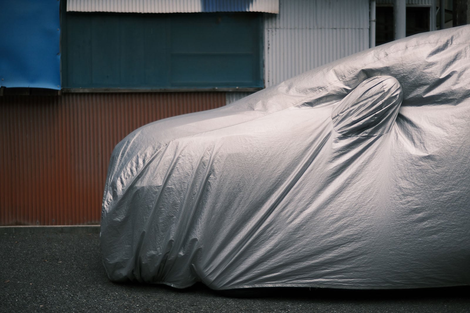 A car covered in a silver protective tarp is parked on asphalt in front of a building with blue and corrugated metal walls.