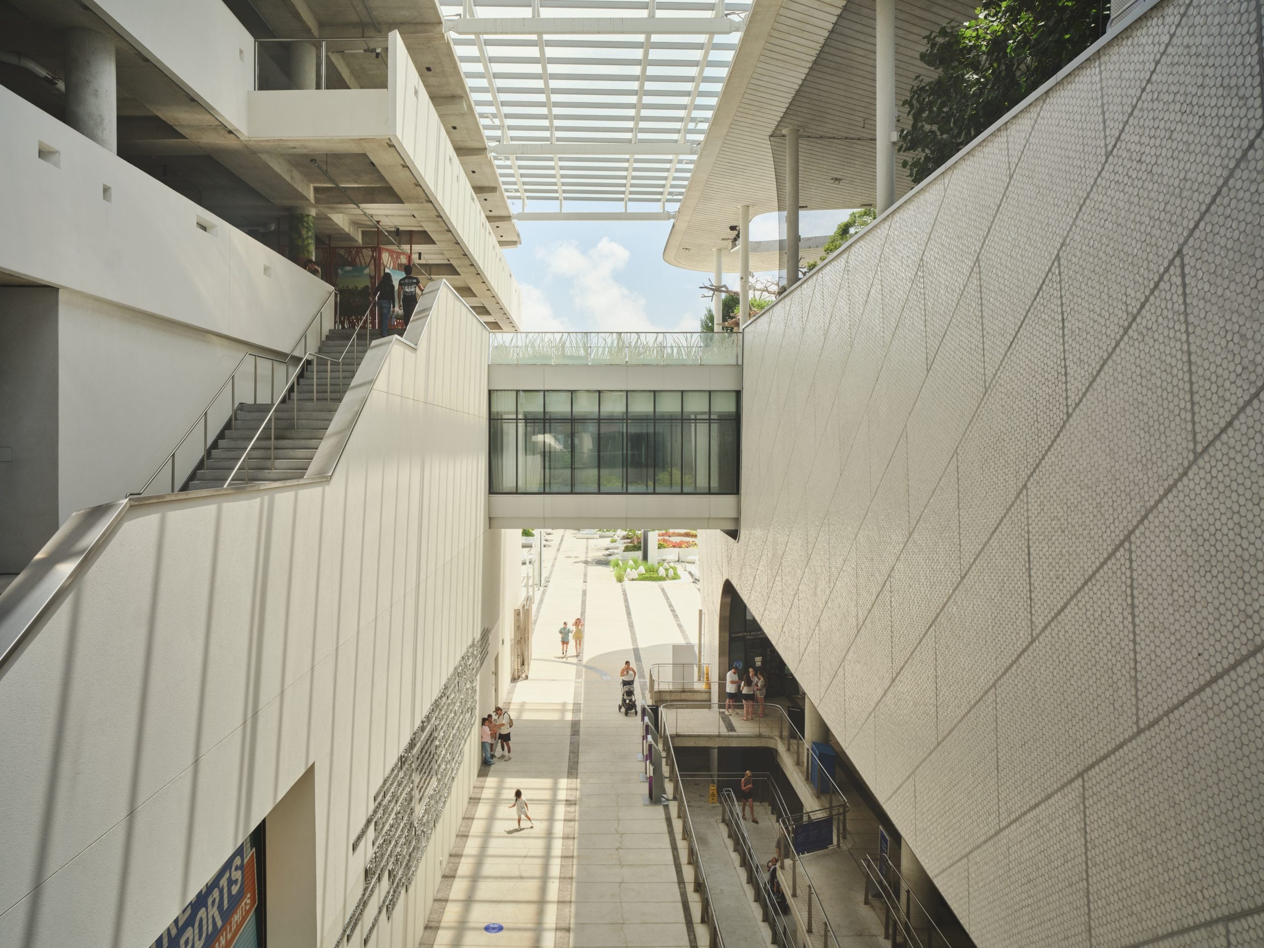 A modern outdoor plaza features a glass-enclosed pedestrian bridge connecting two white, multi-level buildings under a slatted roof structure.