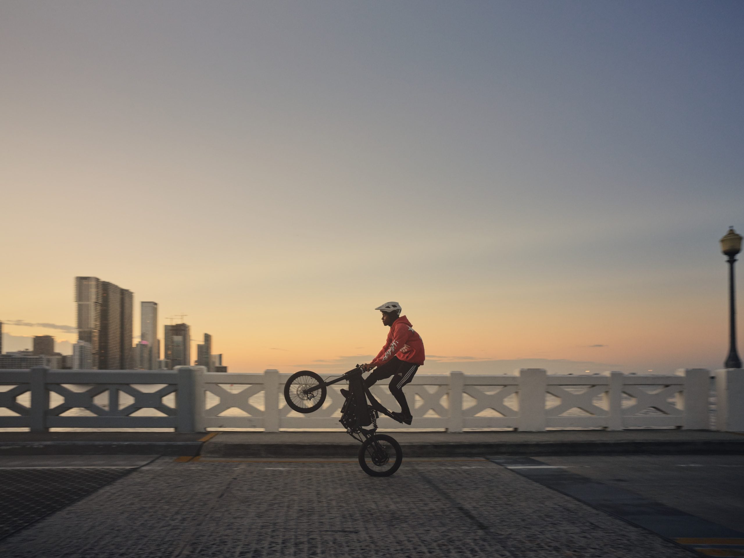 A person wearing a helmet and red hoodie performs a wheelie on an electric bicycle on a bridge at sunset with a city skyline in the background.