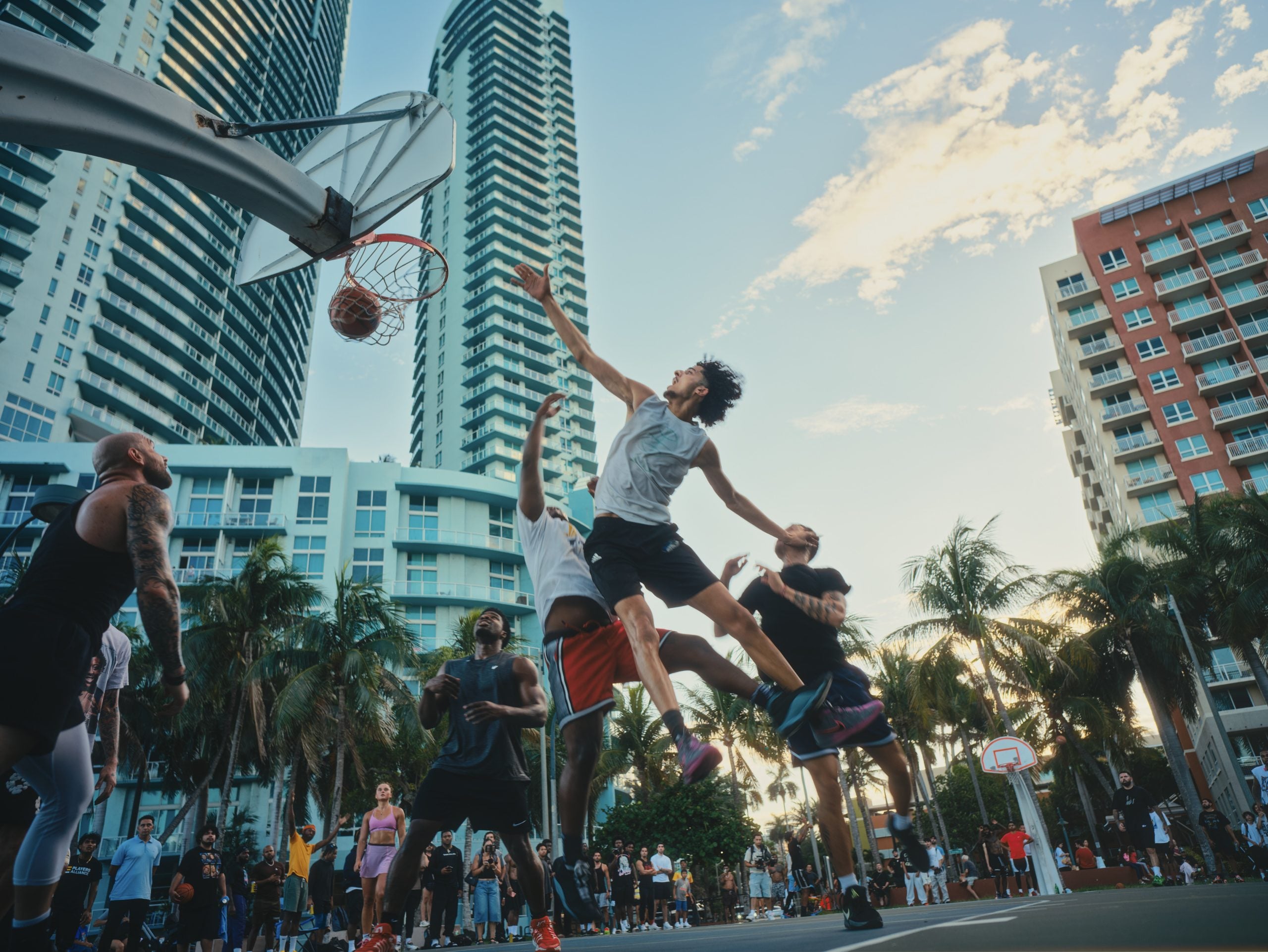A basketball player in a white tank top jumps to block a shot near the hoop, surrounded by other players and spectators on an outdoor court.