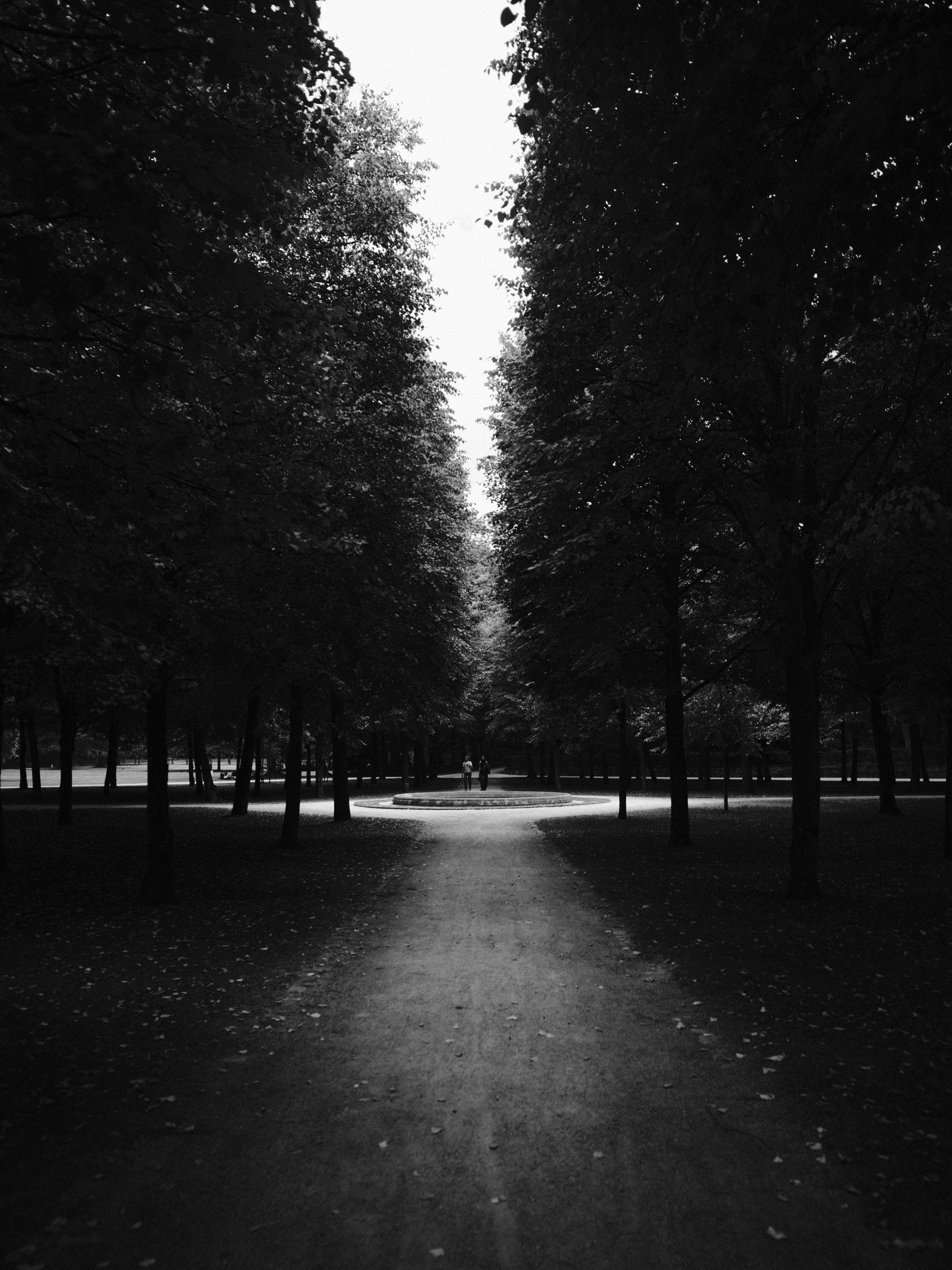 A dirt path leads through a dark, tree-lined park toward a circular fountain where two people stand in the distance.