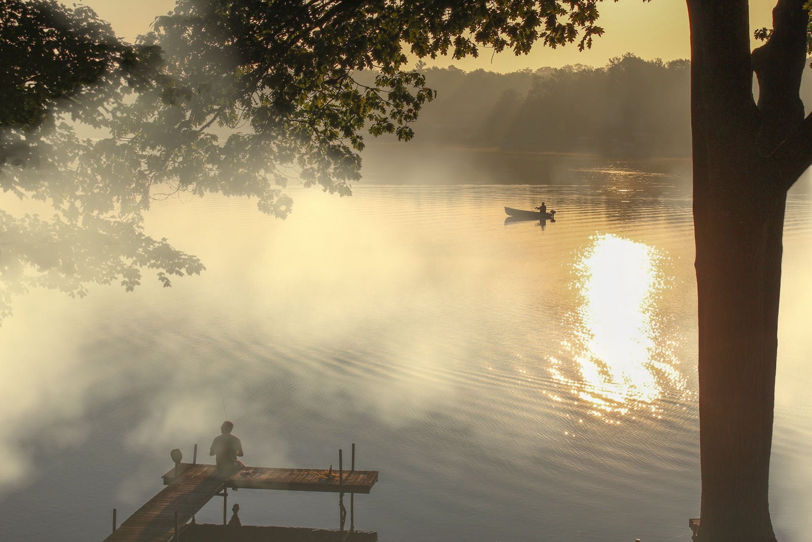 A person sits fishing on a wooden dock at sunrise, while another person in a small boat drifts on the misty lake.