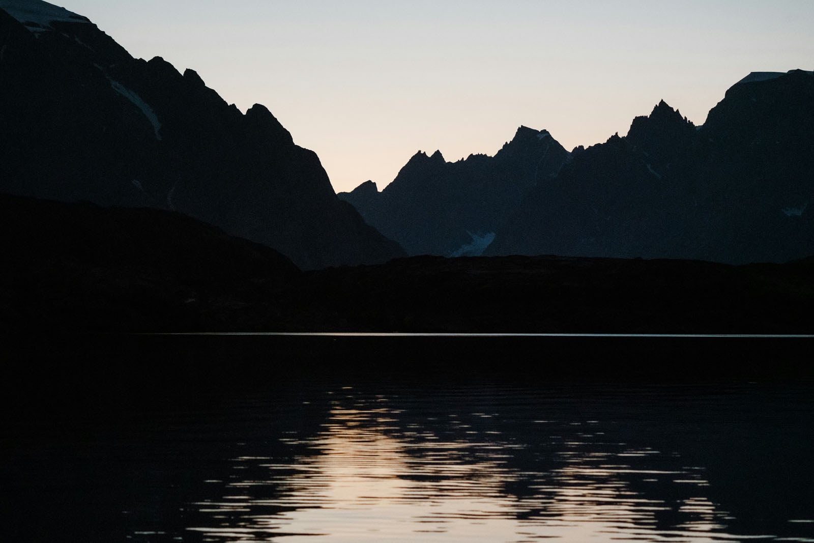 A dark silhouette of jagged mountain peaks rises above a calm lake, reflecting the soft light of a twilight sky.