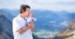 A man in a white polo shirt kisses the forehead of a newborn baby he is holding against a backdrop of mountain ranges.