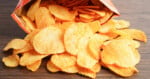 A bag of ridged potato chips lies on its side on a wooden surface, with many chips spilled out in front of it.