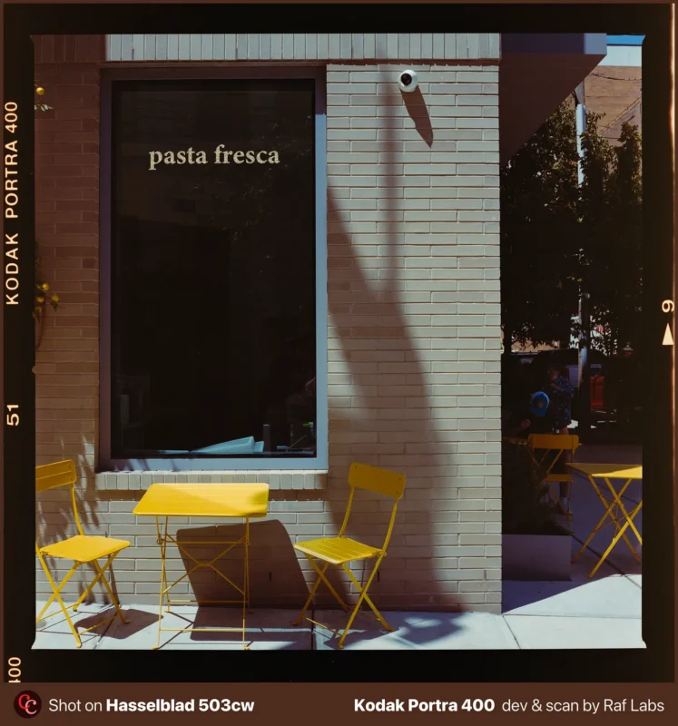 Two yellow metal chairs and a matching table sit on a sidewalk in front of a brick building with a window labeled pasta fresca.