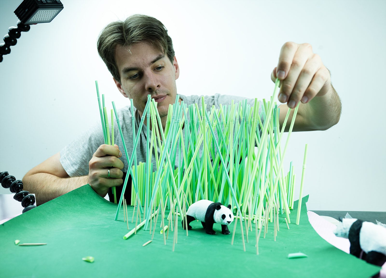 A man carefully places green plastic straws into a green base to create a forest scene for a small panda figurine.
