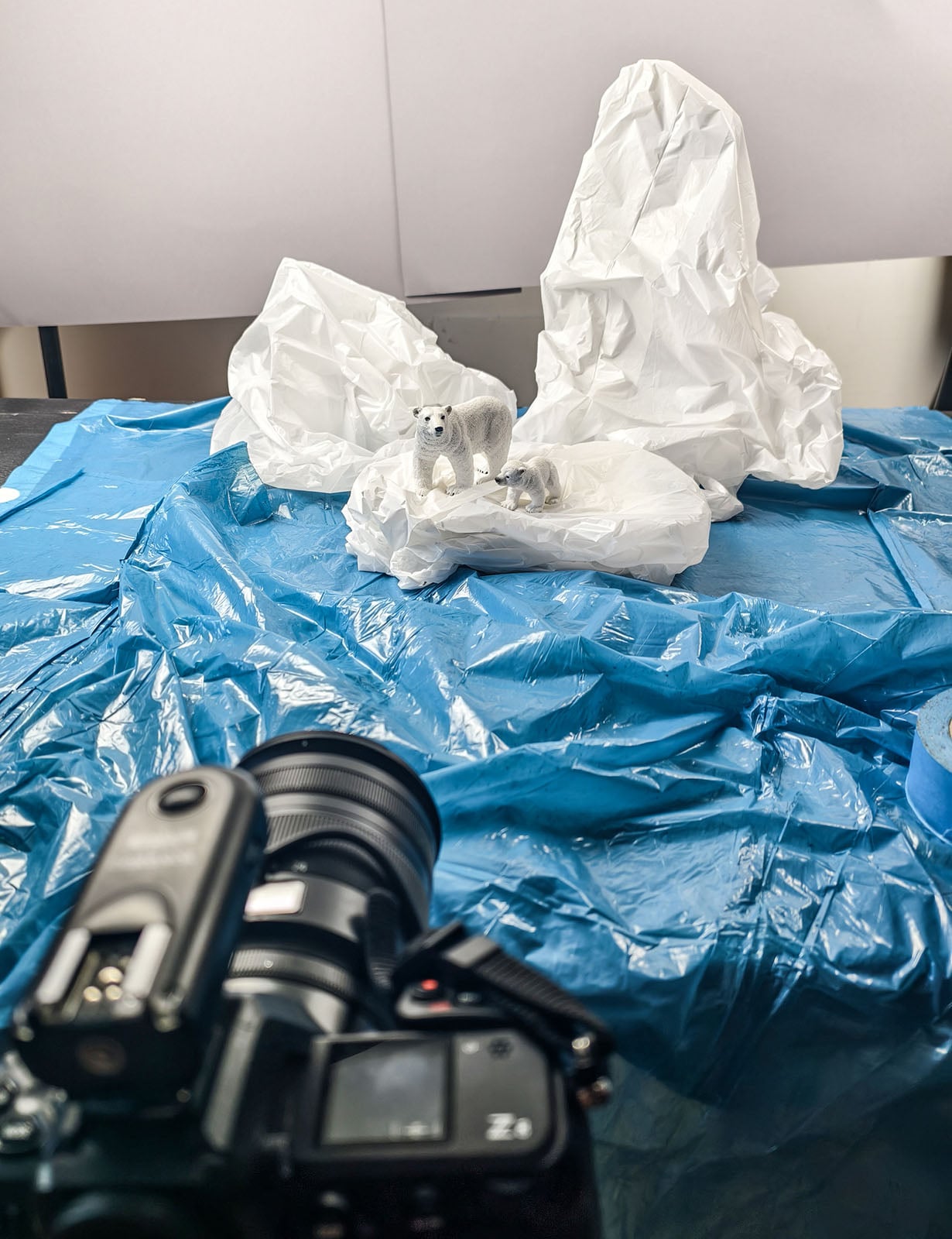 A camera in the foreground is pointed at a diorama featuring two polar bear figurines on crumpled white paper icebergs set against a blue plastic sheet.