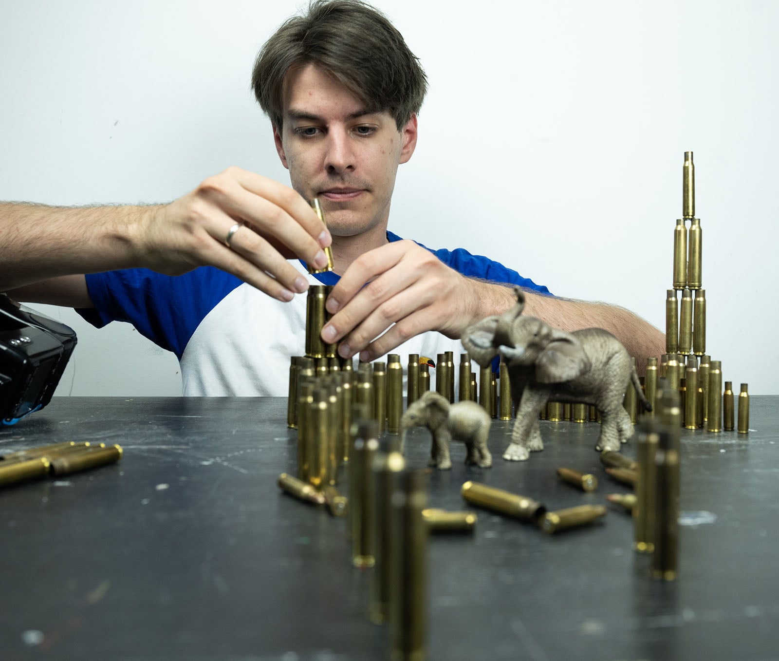A man in a blue and white shirt carefully stacks brass bullet casings on a table next to two toy elephants.
