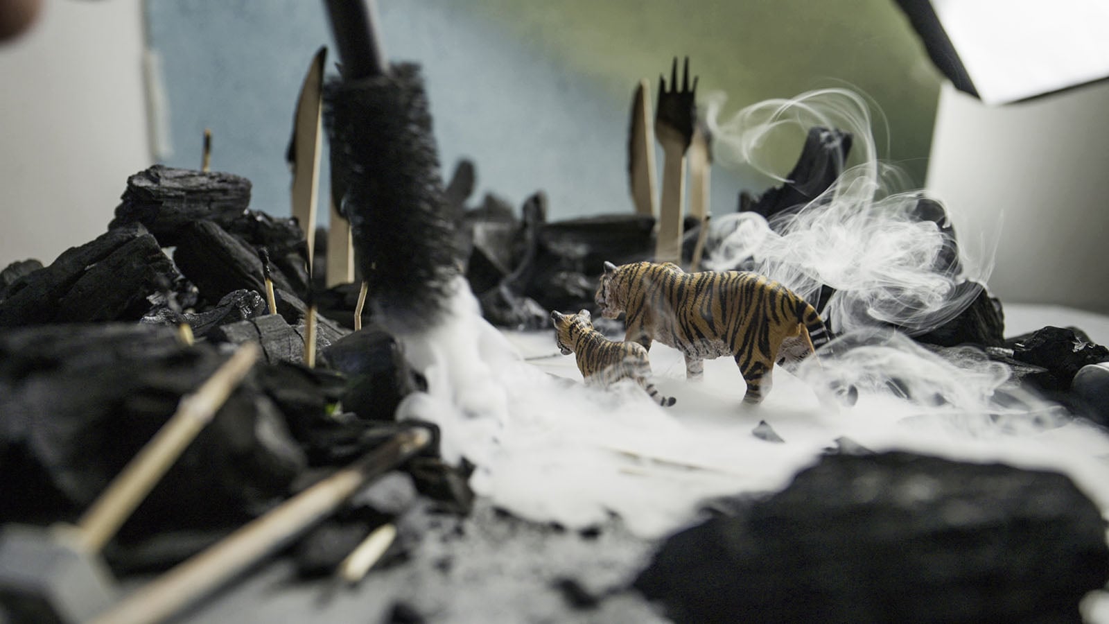 Two toy tigers walk through a landscape of charcoal pieces, wooden utensils, and swirling white smoke.