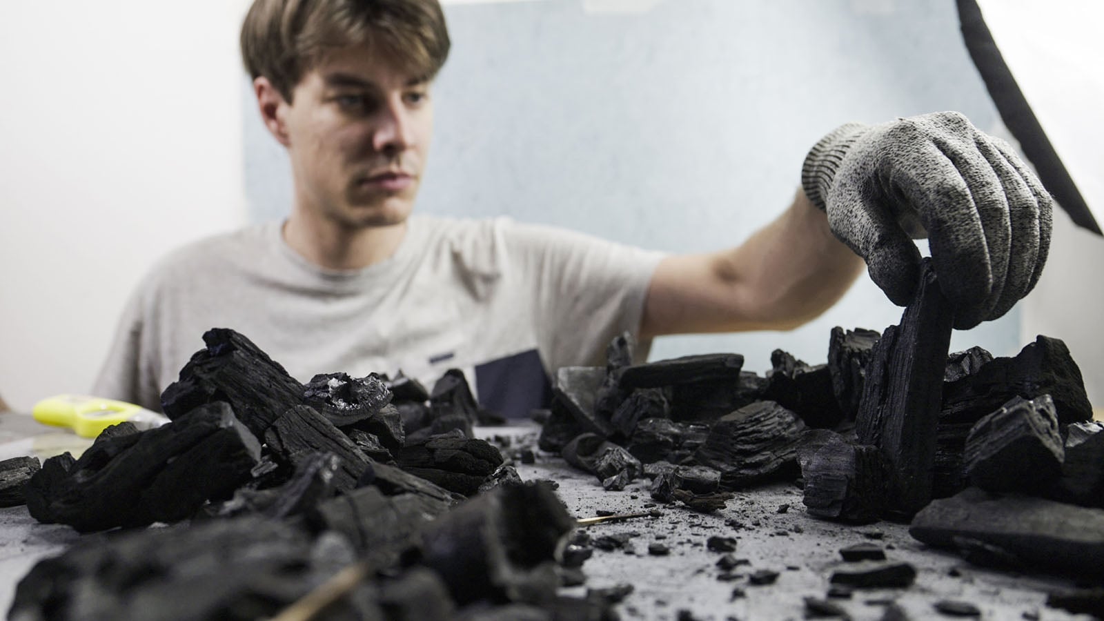 A person wearing a gray work glove reaches out to place a piece of charcoal onto a pile of charcoal fragments on a table.