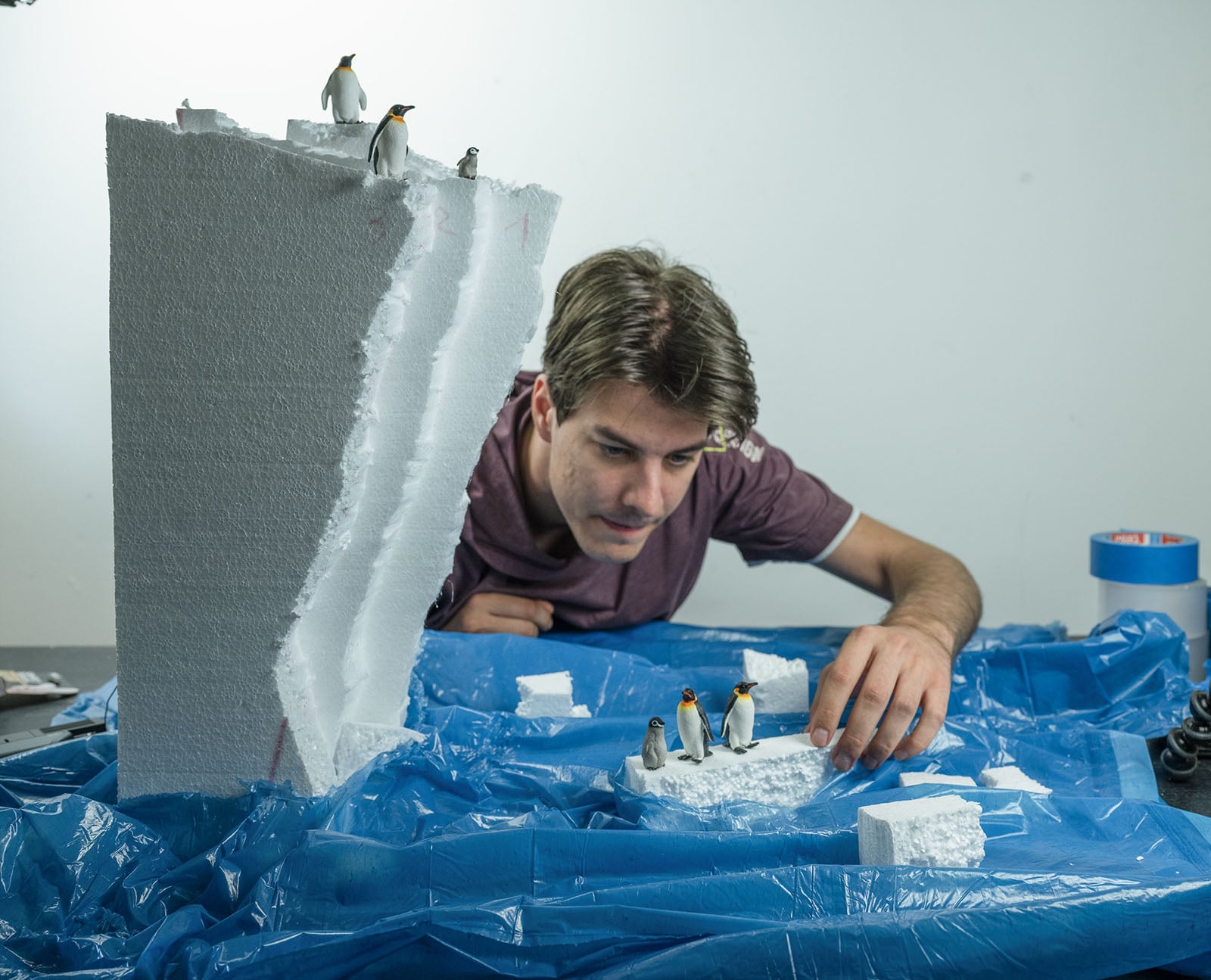 A man leans over a table covered in blue plastic, arranging small penguin figurines on pieces of white foam that represent icebergs.