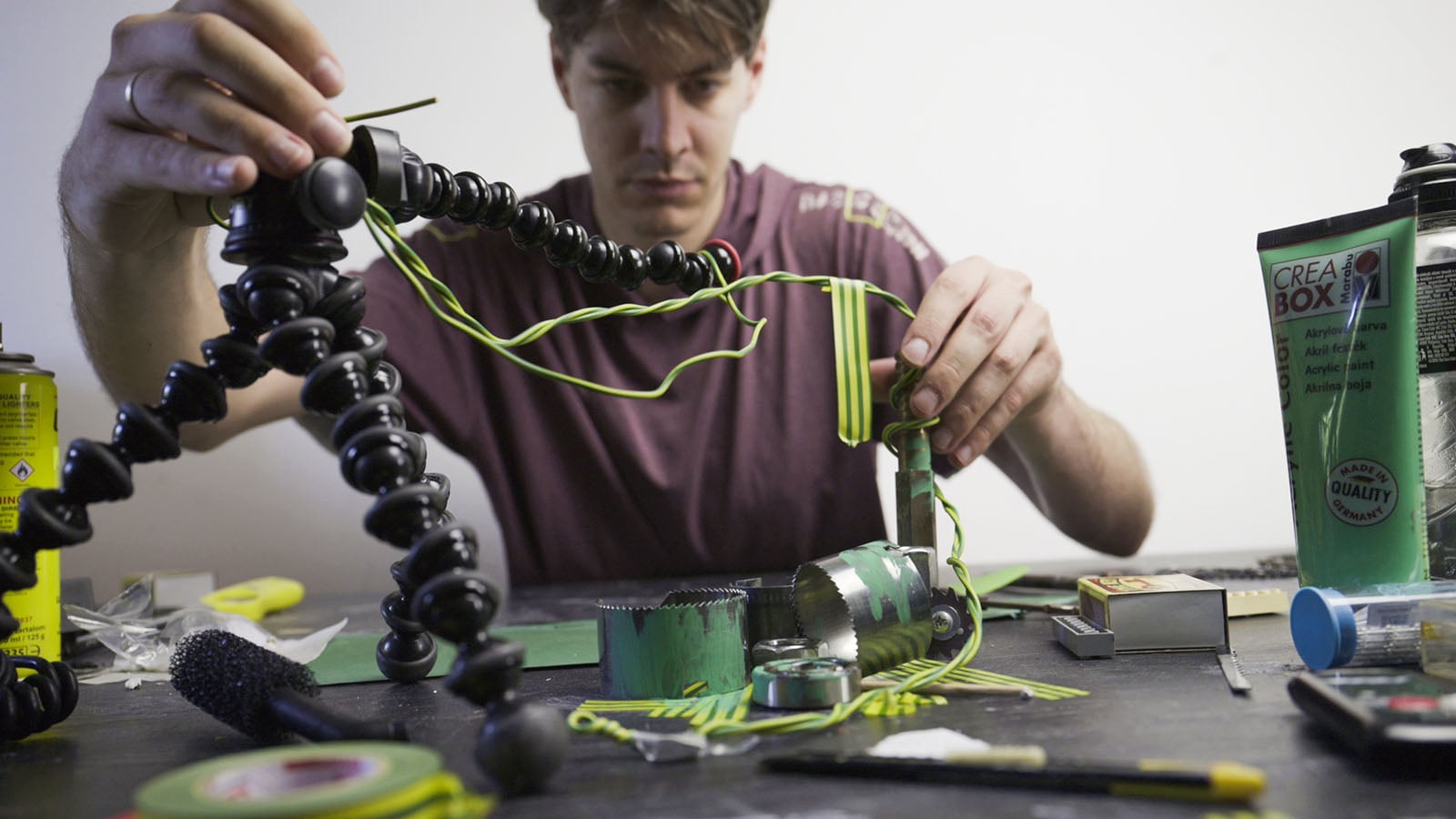 A person sits at a table assembling a project with green and yellow wires, a flexible tripod, and various metal components.