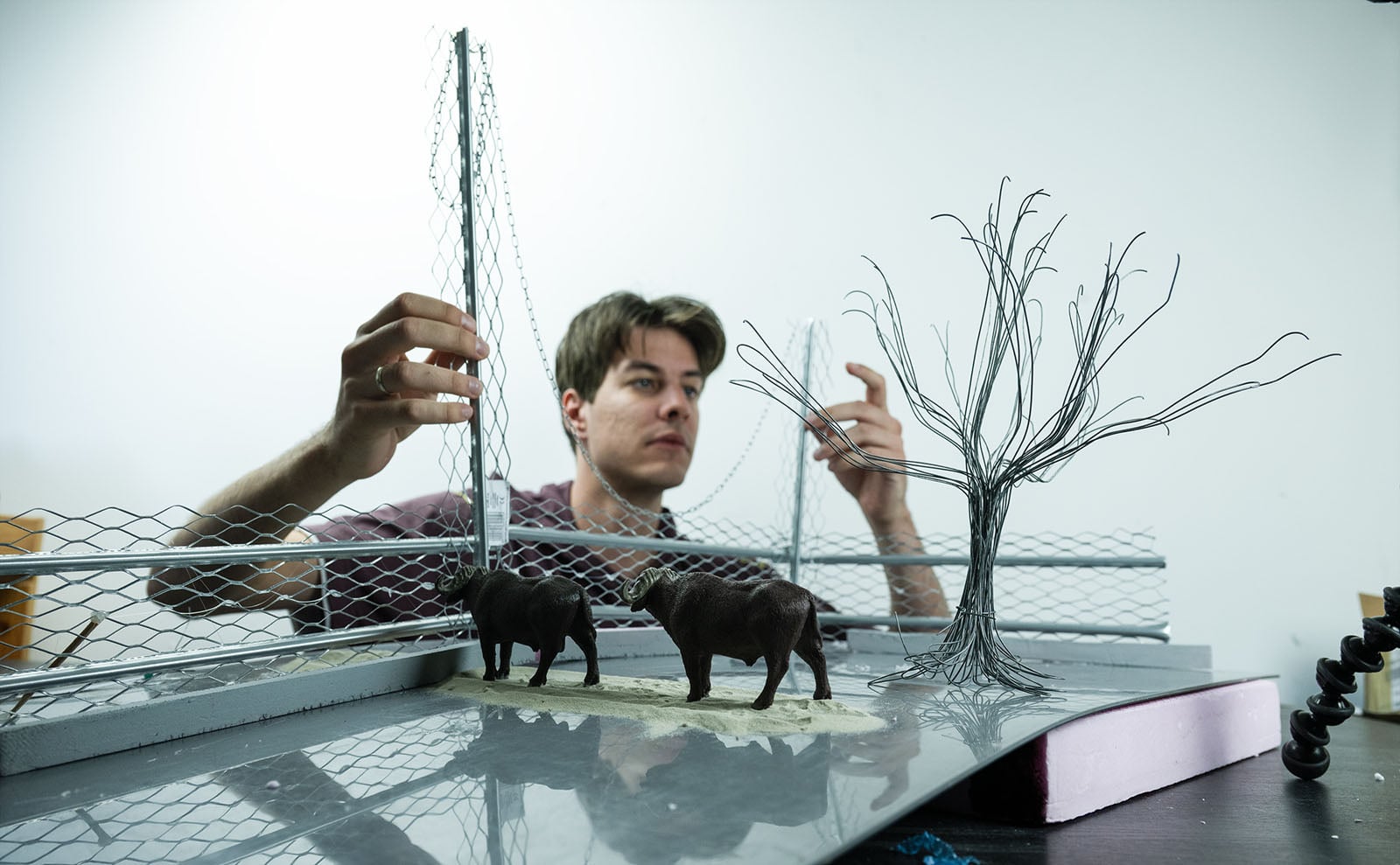 A person adjusts a miniature wire fence and a wire tree model on a platform featuring two small buffalo figurines.