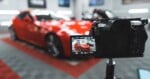 A camera on a tripod records a red sports car parked in a garage, with the car's image displayed on the camera's screen.