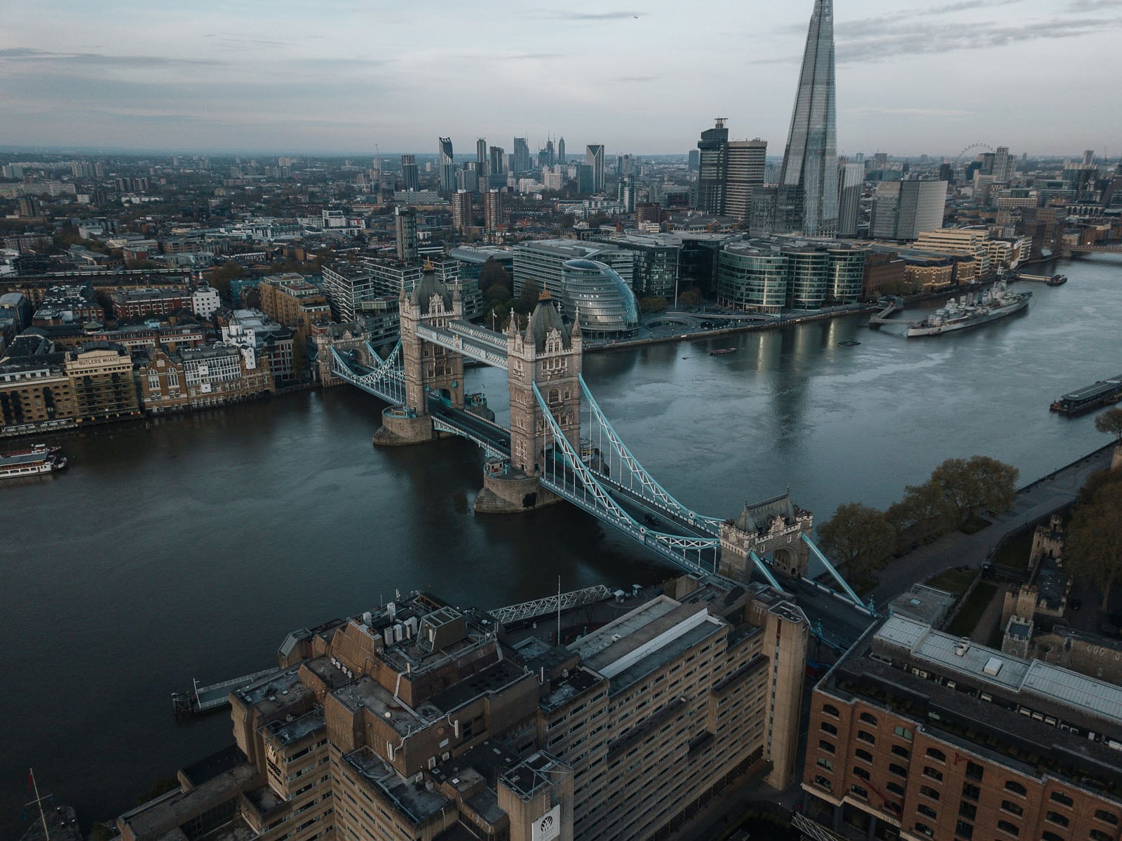 An aerial view of Tower Bridge spanning the River Thames in London, with the Shard and city skyline visible in the background.
