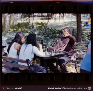 Two women sit at a park bench playing chess against a man who is holding his hand up to his face.