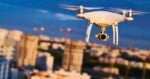 A white quadcopter drone with a camera gimbal flies in front of a blurred city skyline at dusk.