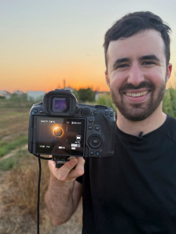 A smiling man holds a camera in front of him, displaying a photo of a solar eclipse on the digital screen.