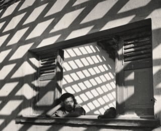 A woman rests her head on her hand while looking out of a window, framed by the geometric shadows of a trellis.