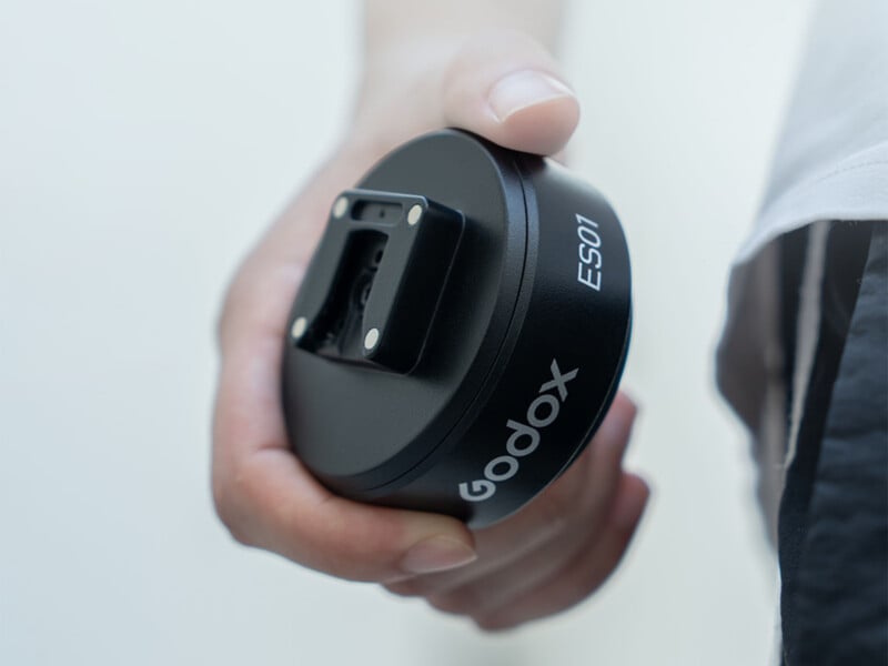 A hand holds a black, circular Godox ES01 device featuring a square mounting bracket on its flat surface.