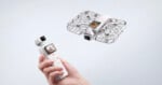 A hand holds a white handheld gimbal camera with a screen, while a matching white drone with a protective cage flies in the air nearby.