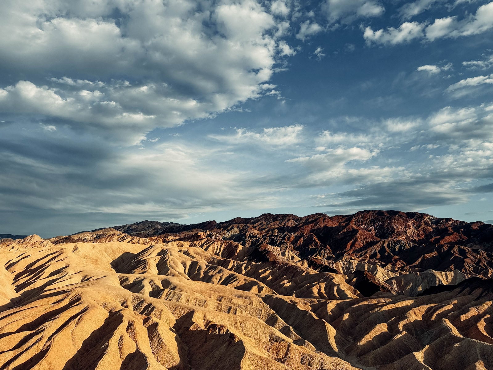 Golden, eroded badlands stretch toward a range of dark, rugged mountains under a blue sky filled with scattered clouds.