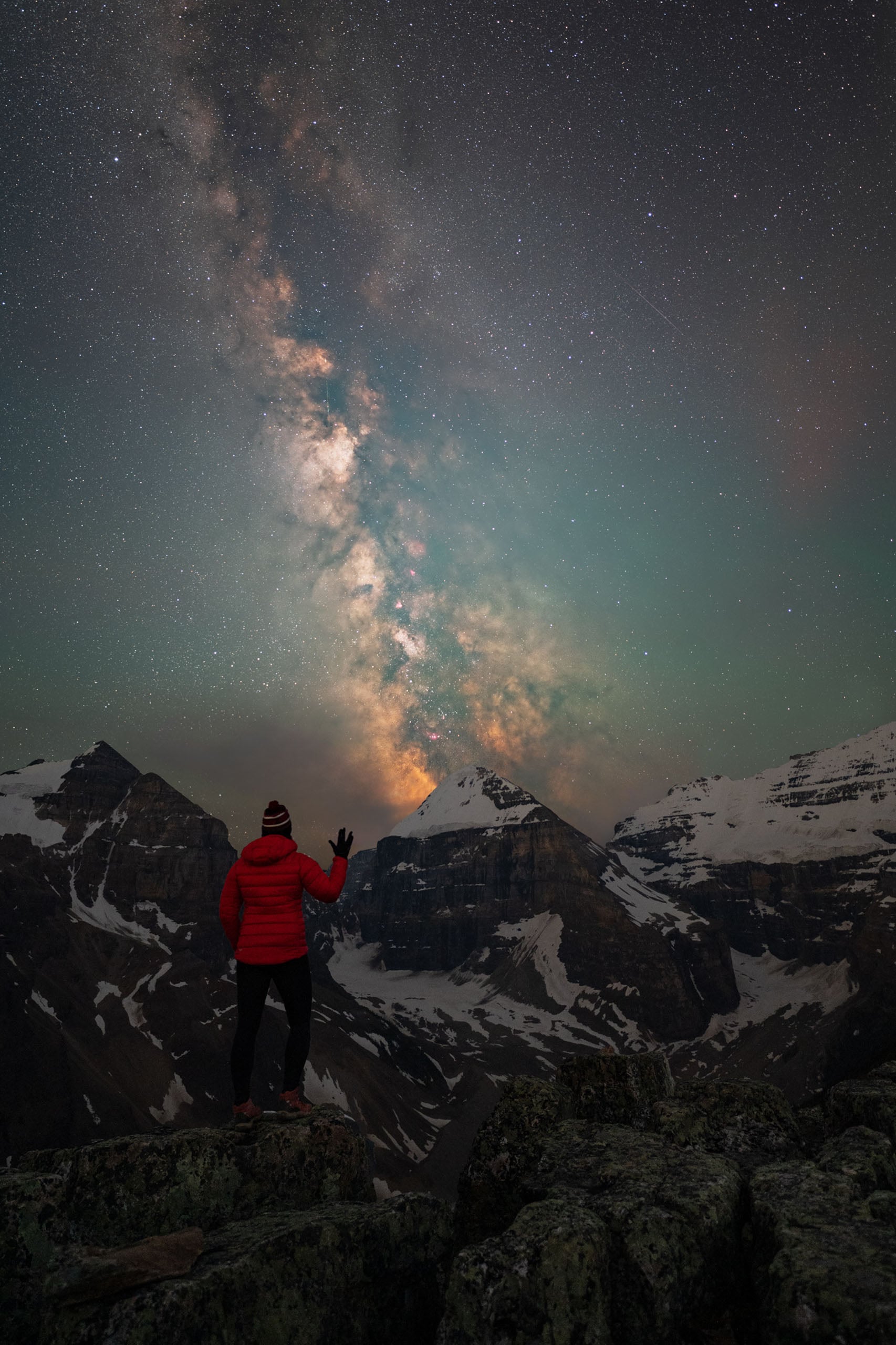 A person in a red jacket stands on a rocky mountain peak at night, gesturing toward the bright Milky Way galaxy visible in the starry sky above snow-capped mountains.
