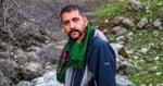 A man with a dark beard and short hair wears a green patterned scarf and a dark blue jacket, standing outdoors in a rocky, natural landscape.