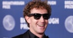 Mark Zuckerberg with curly hair and sunglasses smiles while wearing a black suit against a blue Breakthrough Prize backdrop.
