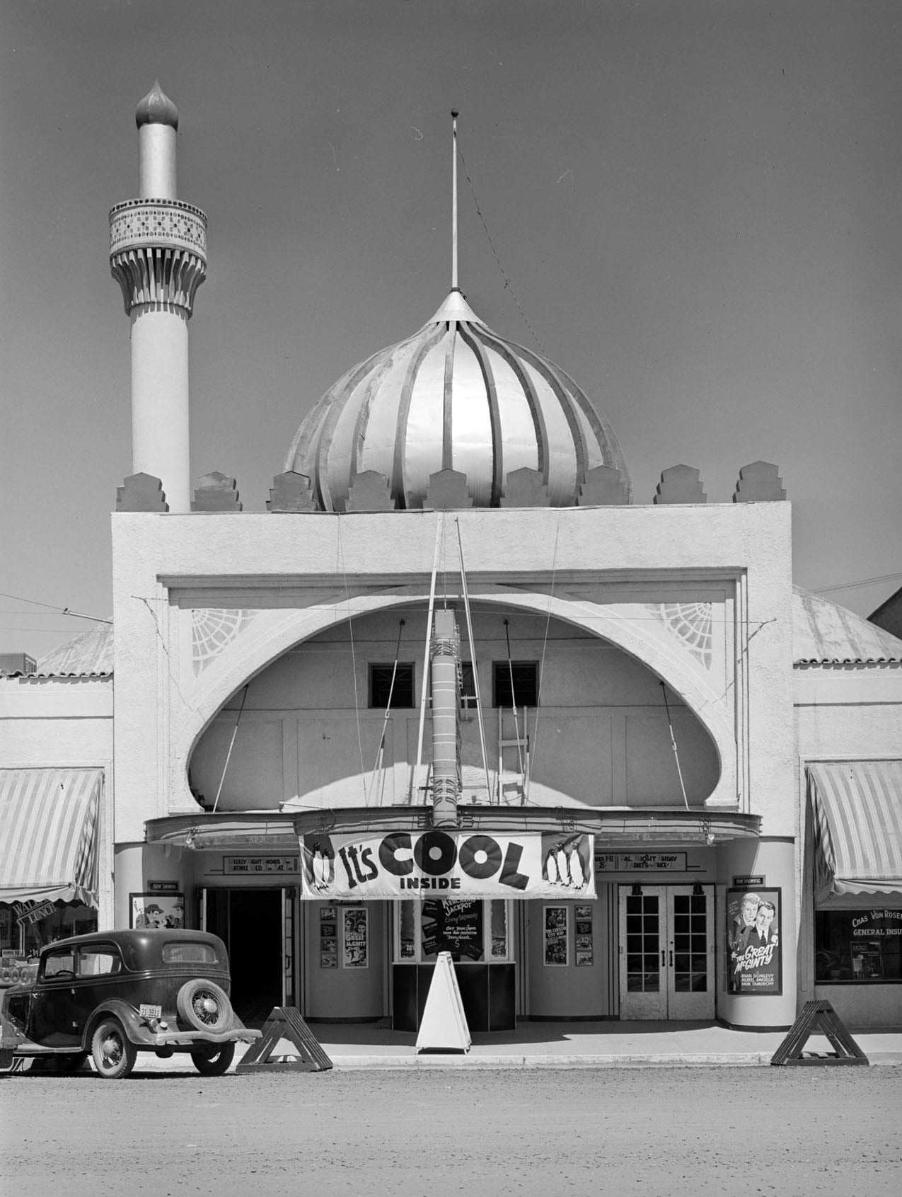A black-and-white eye-level shot of a theater with a dome and minaret, featuring a banner reading "It's Cool Inside" and a vintage car parked in front.